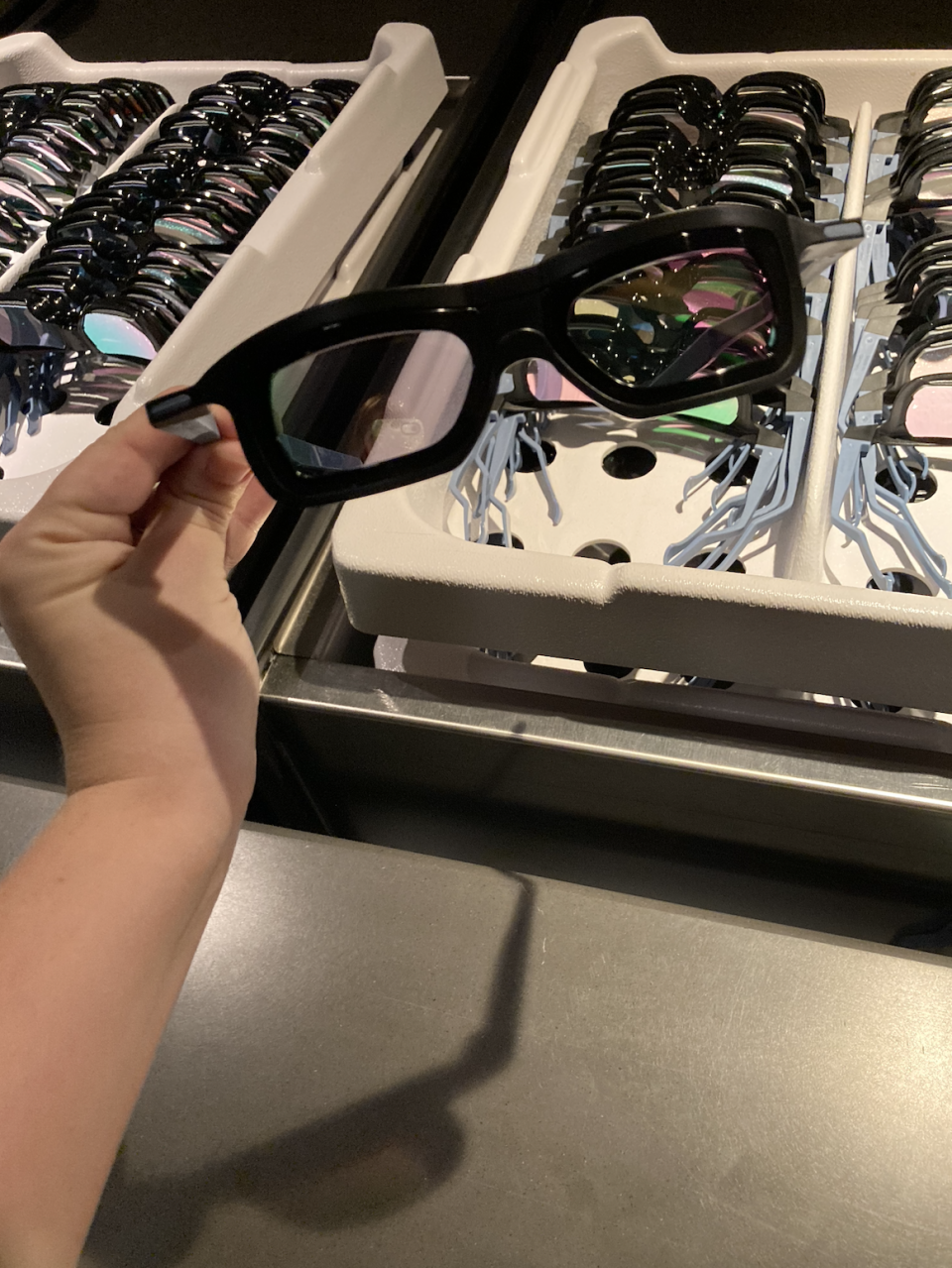 A person holds a pair of black 3D glasses in front of a tray filled with many other similar 3D glasses