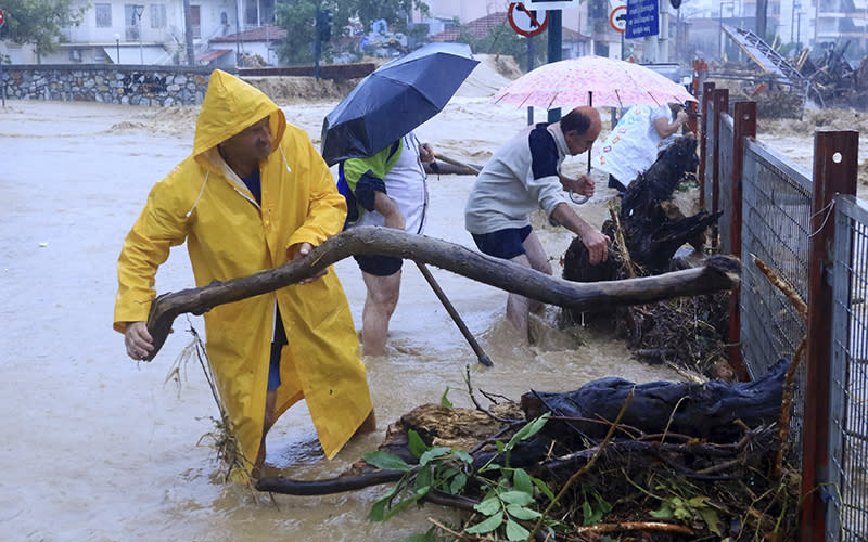 Residents remove debris from a flooded area. One man is wearing a yellow hooded rain jacket as he pulls a branch away from a fence. Behind him are several other people with umbrellas