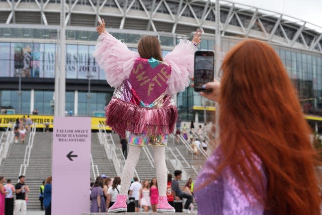 A woman taking a photo of a young girl with her arms in the air outside Wembley Stadium while wearing a costume that has the word Swiftie on the back