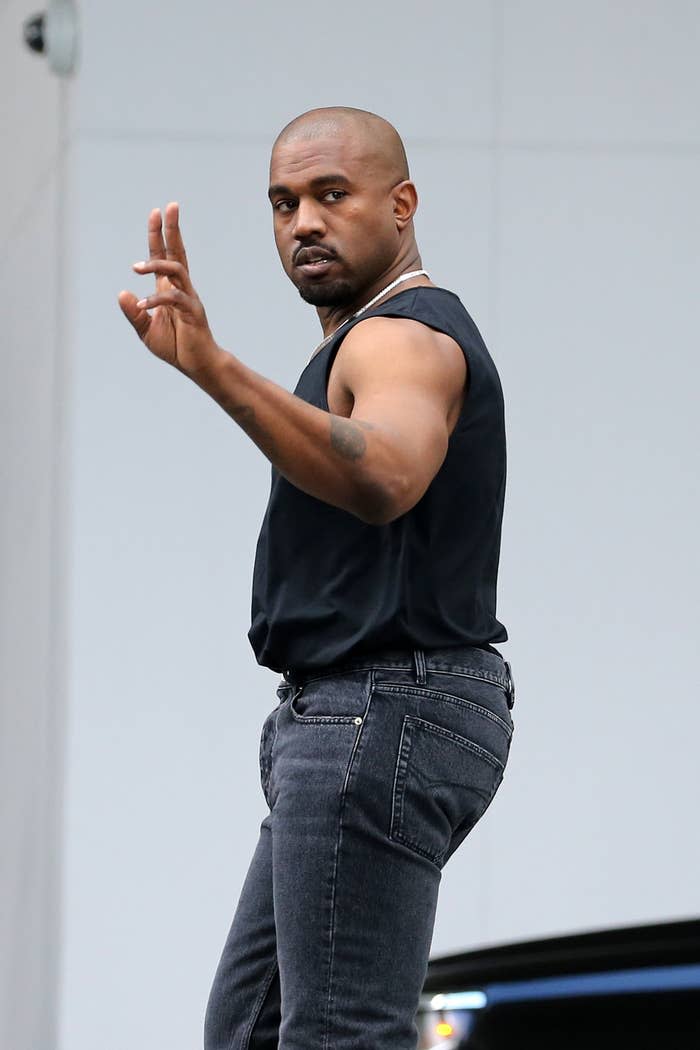 Kanye walking outside in a sleeveless shirt and jeans as he waves at somebody