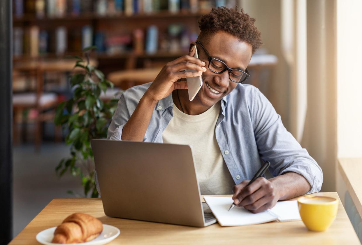 Cheerful black guy talking on smartphone while studying online at coffee shop