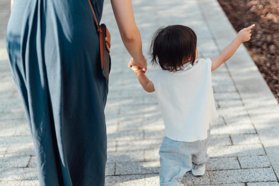 An adult holds hands with a child, who is pointing forward as they walk on a paved path