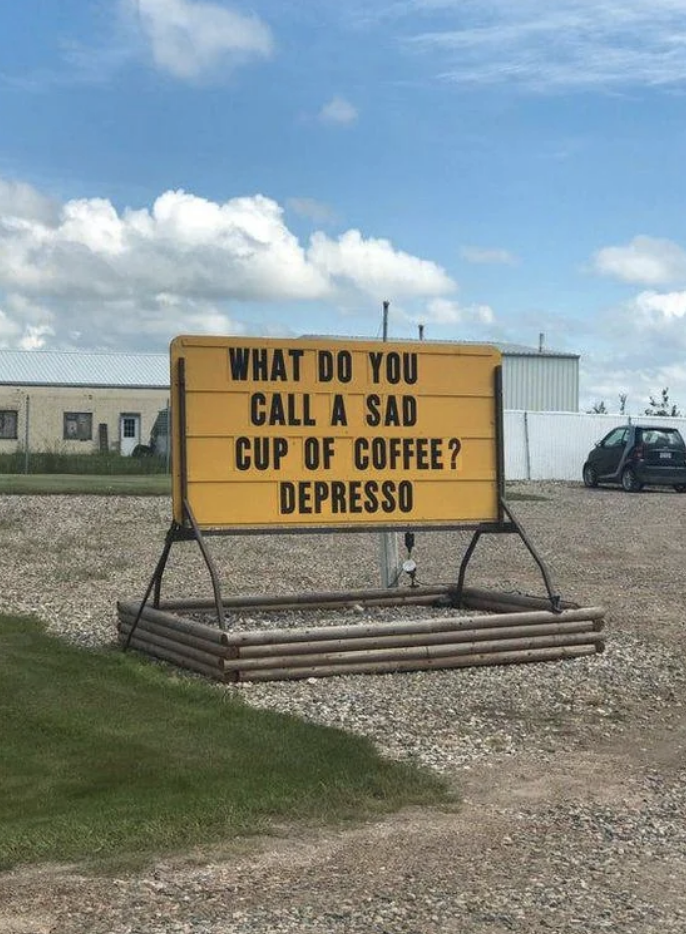 Sign with joke: "WHAT DO YOU CALL A SAD CUP OF COFFEE? DEPRESSO" displayed outside, with a grass field and building in the background