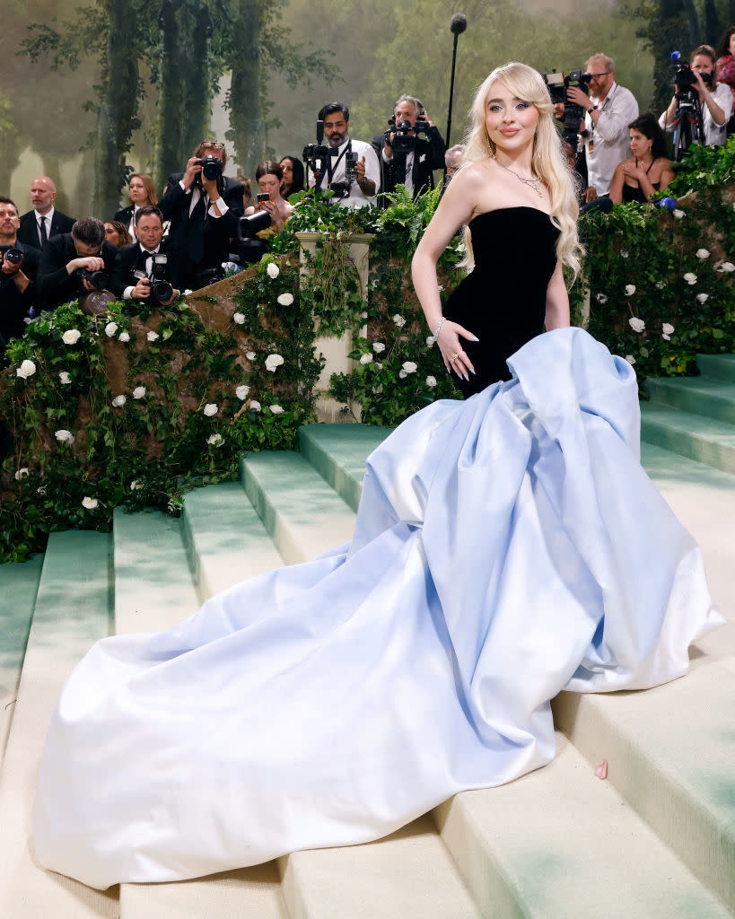 Woman in elegant gown with large blue bow at an event with photographers in background