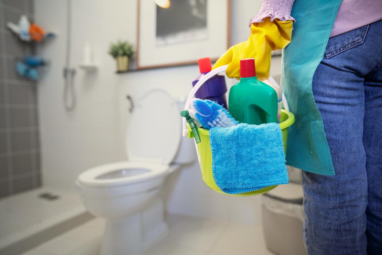 The cleaning woman is standing in the bathroom holding a blue bucket full of chemicals and cleaning utensils