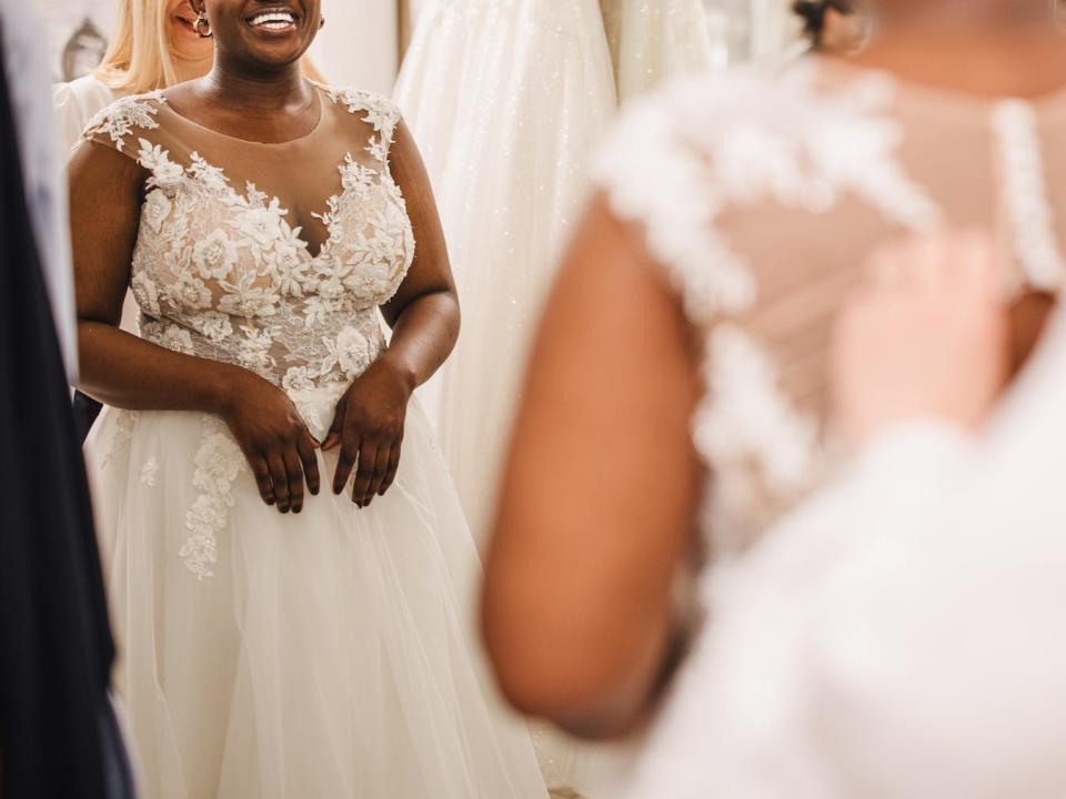 A bride smiles as someone adjusts a wedding dress for her in a bridal salon.