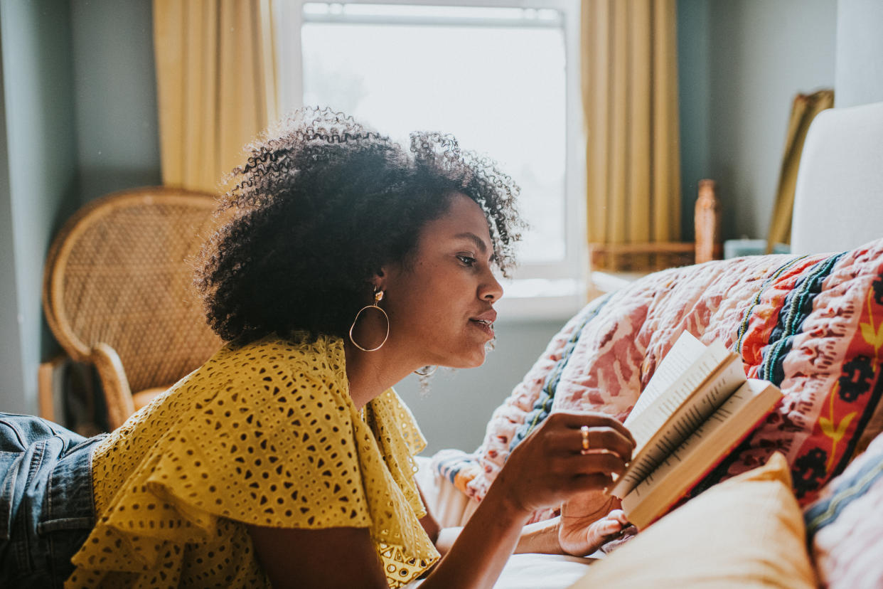 A beautiful woman lies on her stomach in a comfortable, stylish bedroom. She concentrates on reading her book. Space for copy.