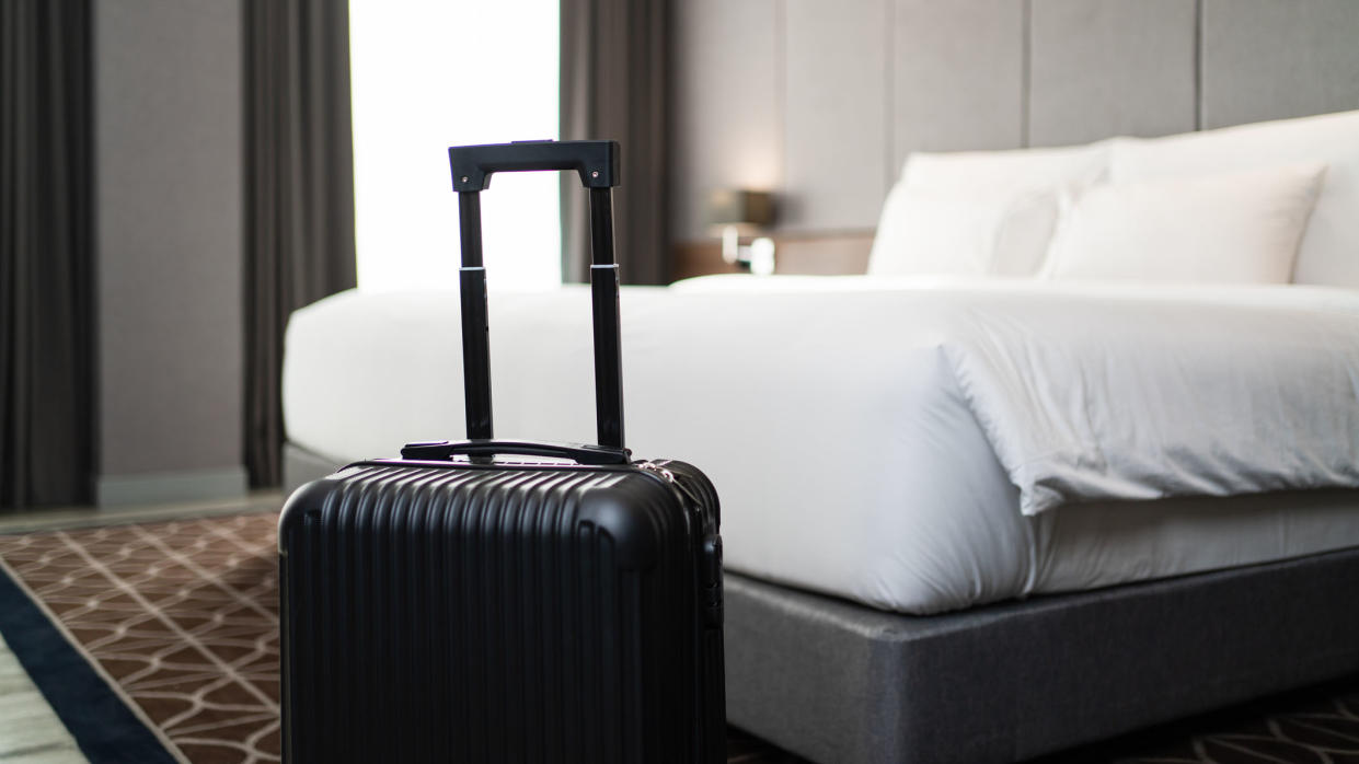  A black suitcase in front of a white bed in a grey hotel room. 