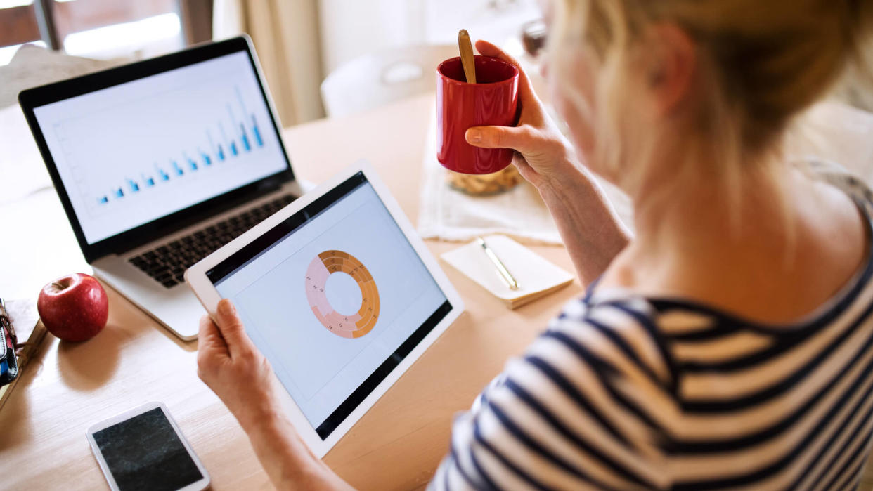 Unrecognizable senior woman with laptop and tablet sitting at ther kitchen table, searching or reading something, holding a cup of coffee.