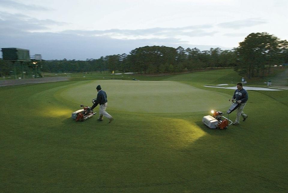 Two workers mow a golf course green in early morning, using lighted mowers