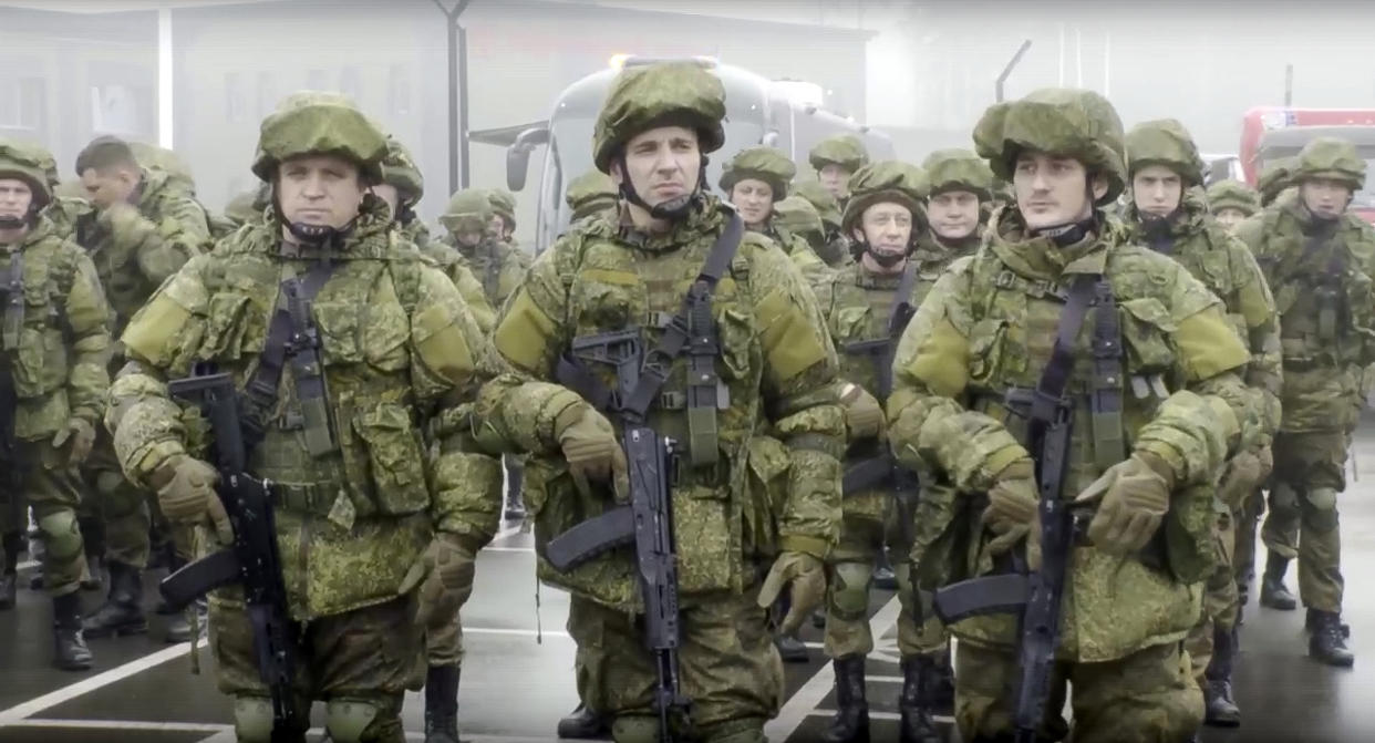 Heavily armed Russian recruiters in green uniforms stand to attention, right hands on the trigger of their firearms, with a phalanx of troops behind them.