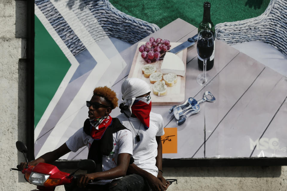 Protesters drive a motorcycle past a billboard advertising a lifestyle few Haitians have access to, in Petion-Ville, Port-au-Prince, Haiti, Sunday, Oct. 13, 2019. Thousands of Haitians joined a largely peaceful protest called by the art community Sunday to demand Moïse resign, increasing pressure on the embattled leader after nearly a month of marches that have shuttered schools and businesses.(AP Photo/Rebecca Blackwell)