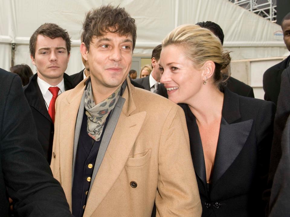 Jamie smiling in a camel coat and Kate resting her chin on his shoulder, wearing a blazer.