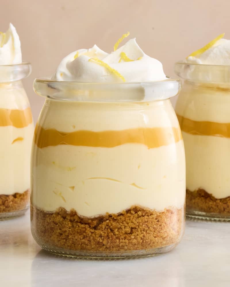 Head on shot of 3 small glass jars of creamy lemon mousse with layers of graham cracker crumbs and whipped cream, and topped with lemon zest.