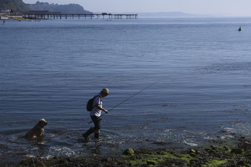 A man is followed by a dog as he moves to another spot to fish in the Teign estuary in Shaldon, Devon, England, Sunday July 18, 2021. (AP Photo/Tony Hicks)