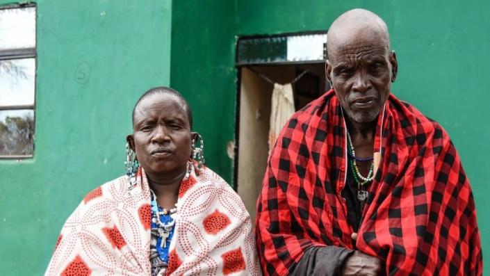 Man and woman both in colourful shawls staring into the camera. They are standing in front of a green house