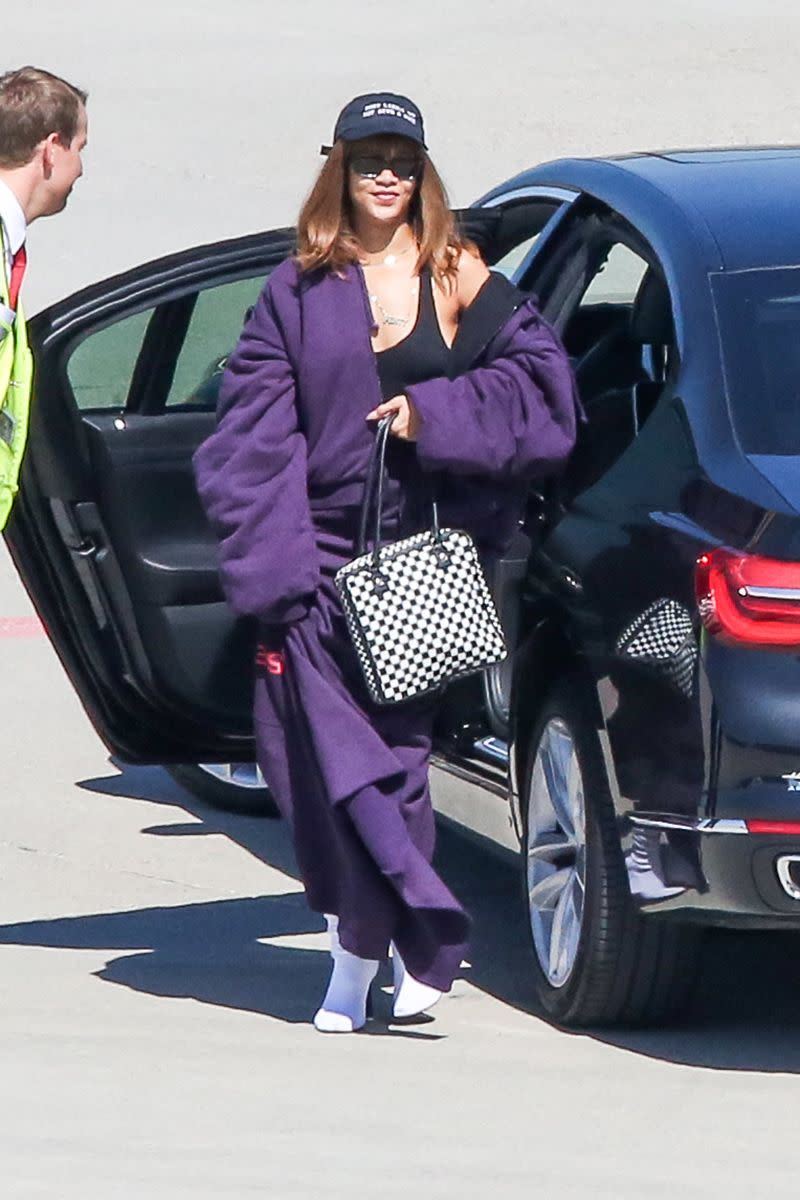 <p>Rihanna heads to her private jet in Zurich cloaked in a purple oversized coat, with a checkered boxy handbag, white boots, a black tanktop, baseball hat and sunglasses.</p>