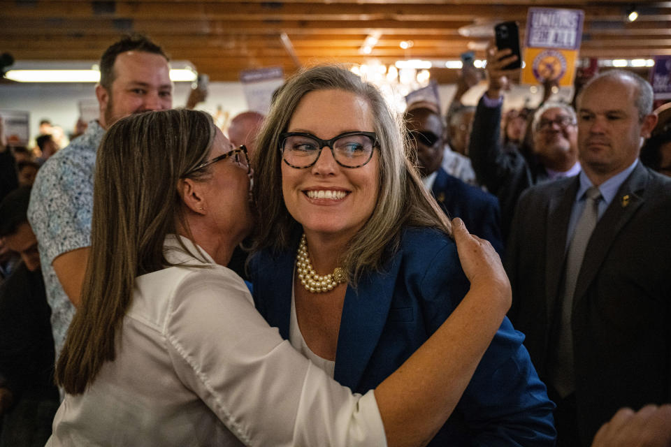 Katie Hobbs, beaming, and wearing glasses and three strings of pearls, hugs her twin sister, Becky Hobbs, as supporters applaud.