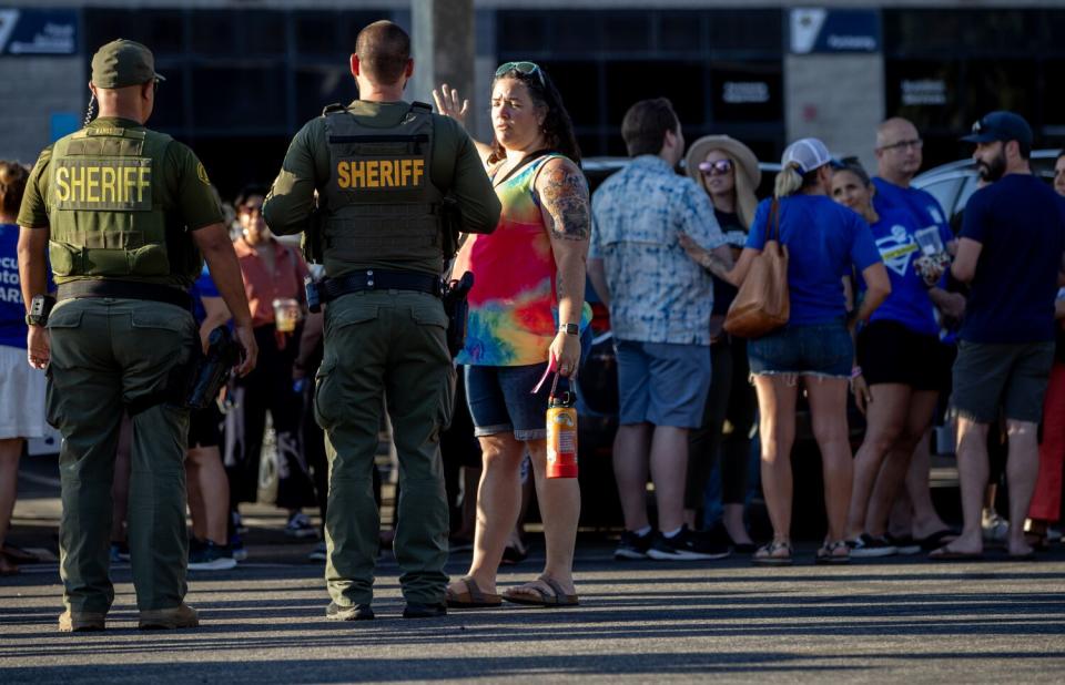 Men in green uniforms labeled "Sheriff" speak with people waiting in the shade outside a building