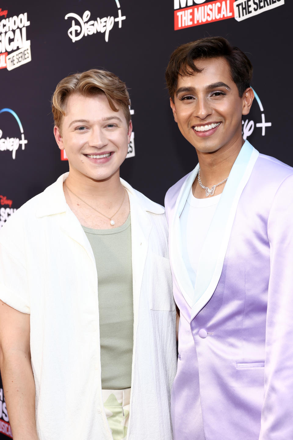Two actors from "High School Musical: The Musical" smiling at a promotional event. One wears a white outfit, the other a lavender suit