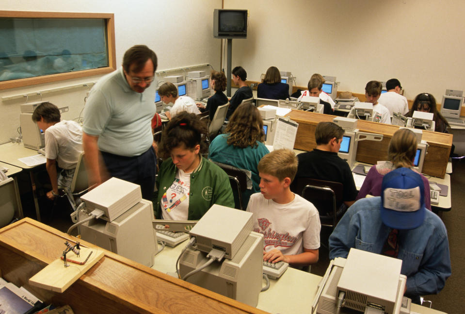 A classroom with students working on computers. A teacher is assisting a student. The room has several desktop computers on desks