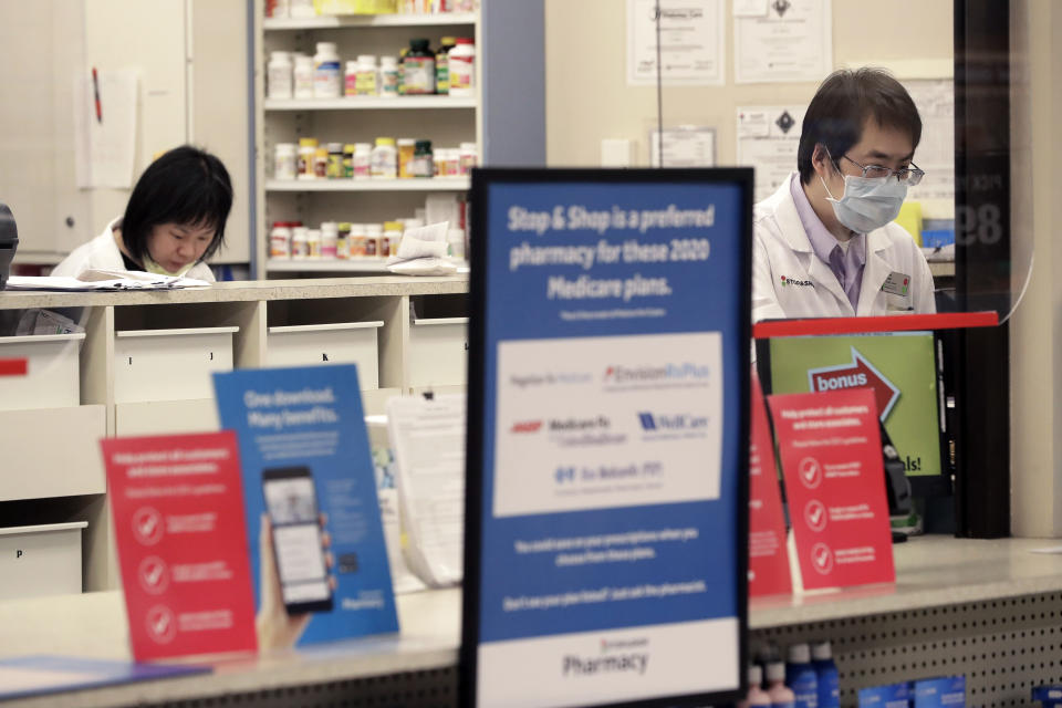 A pharmacist, right, stands behind a plastic shield at a grocery store, Thursday, March 26, 2020, in Quincy, Mass. Grocery stores across the U.S. are installing protective plastic shields at checkouts to help keep cashiers and shoppers from infecting each other with the coronavirus. The new coronavirus causes mild or moderate symptoms for most people, but for some, especially older adults and people with existing health problems, it can cause more severe illness or death. (AP Photo/Steven Senne)