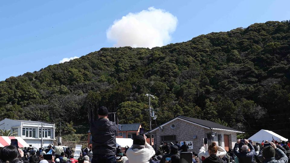 a cloud of smoke rises above a hillside as onlookers point cameras up at it