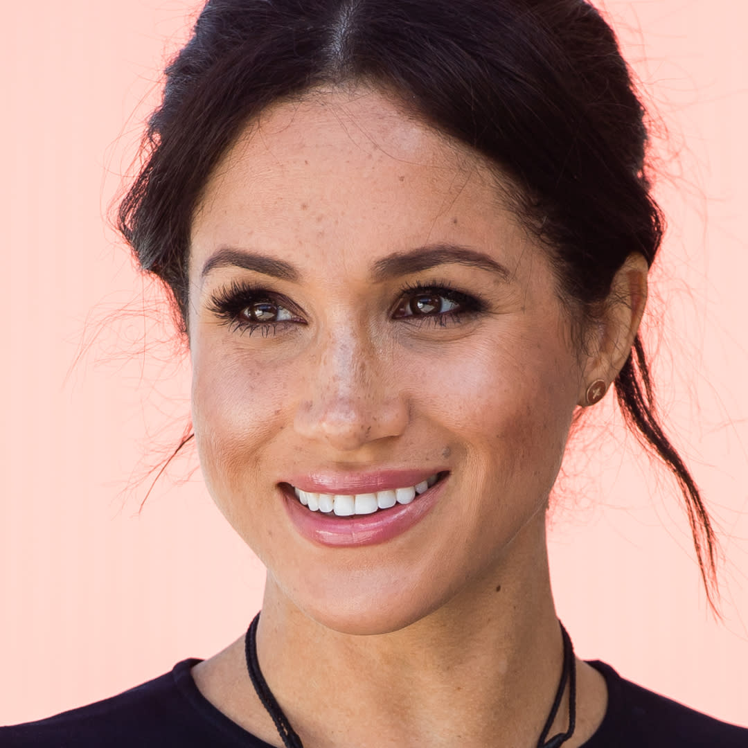 Meghan Markle wearing a black dress with her hair up in front of a plain backdrop. 