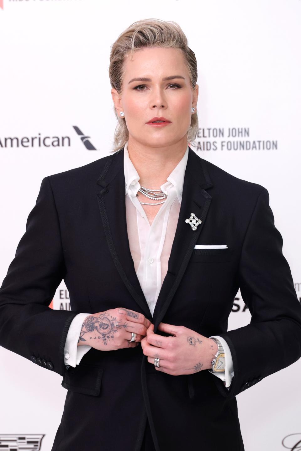 Ashlyn Harris in a tailored black suit with layered necklaces, standing with hands clasped, against a backdrop with logos