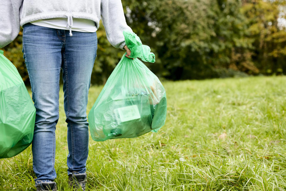 A person is standing in a grassy area holding two green garbage bags, one in each hand. The focus is on the bags and the person's lower body