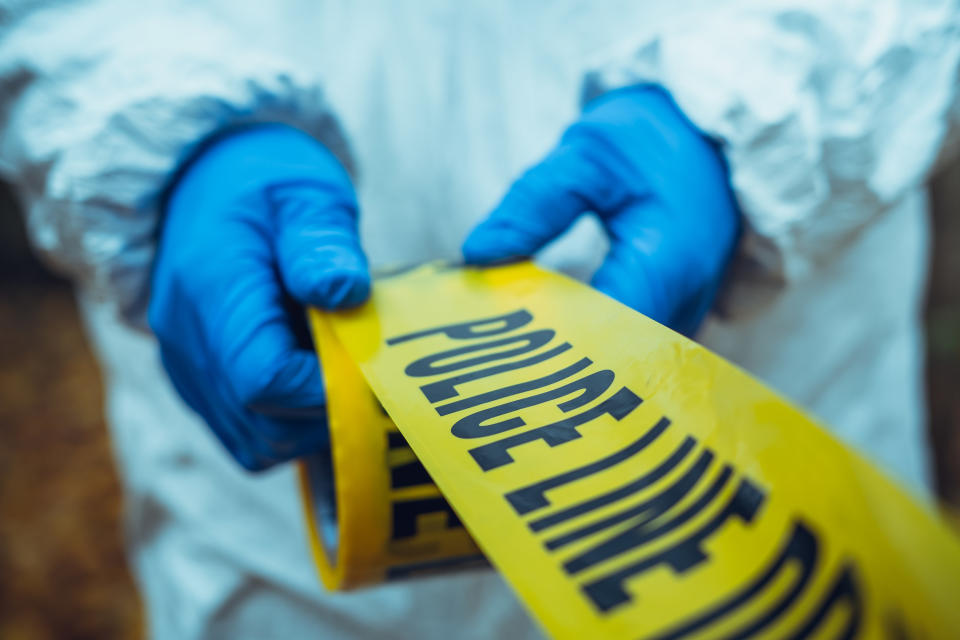 Someone with blue gloves and white suit unrolling yellow police tape