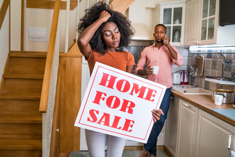 An African American woman, looking frustrated, holds a sign saying Home for Sale in a galley kitchen with her male partner behind her, holding a coffee cup in one hand and conducting a cellphone conversation.