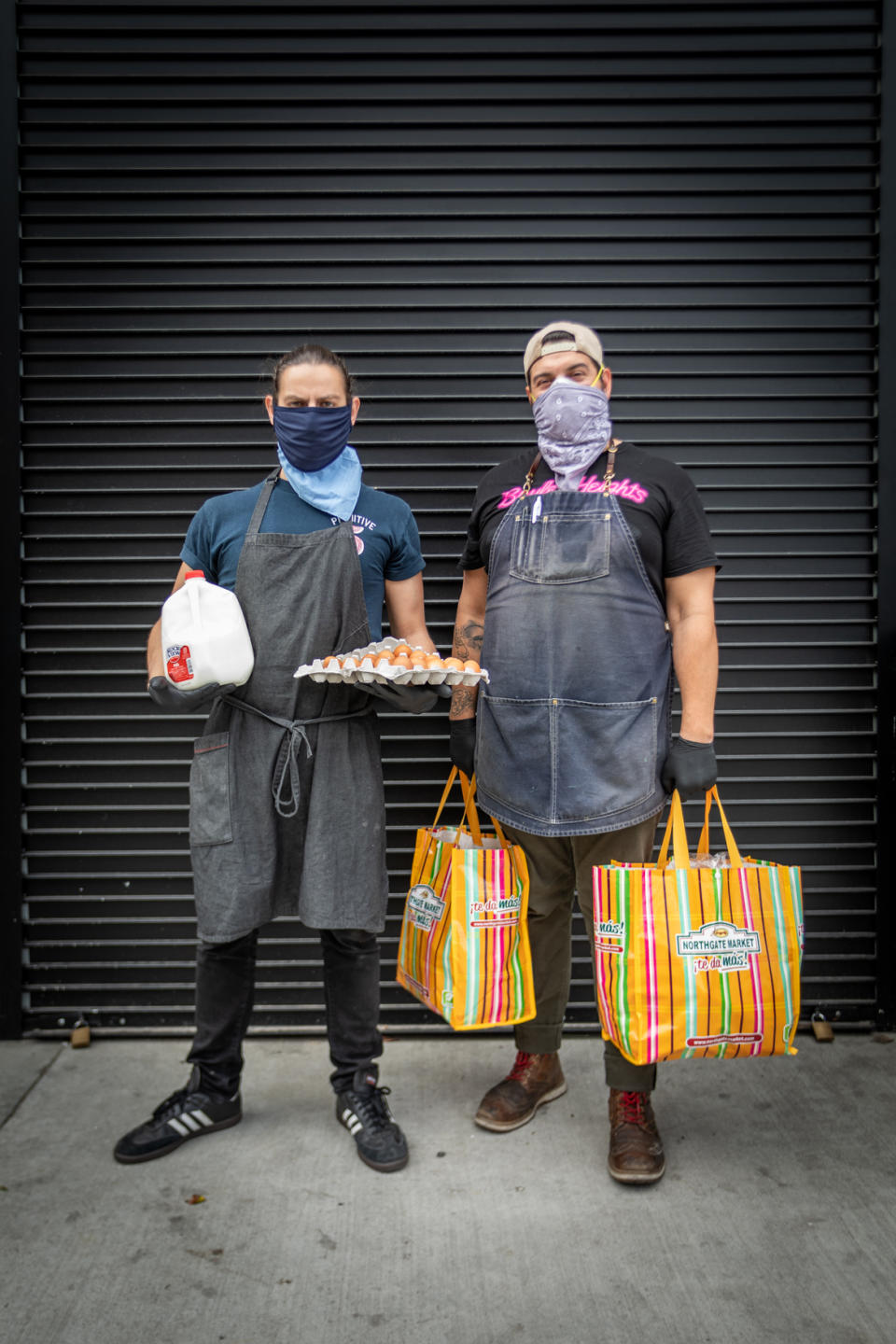 Damian Diaz and Othón Nolasco of No Us Without You stand on the sidewalk wearing face masks and carrying groceries.