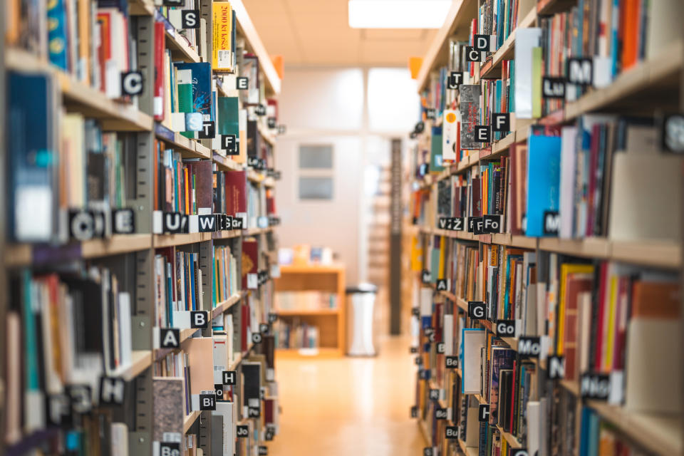 A library aisle with neatly arranged books on shelves labeled with letters and sections, leading to a brightly lit seating area in the background