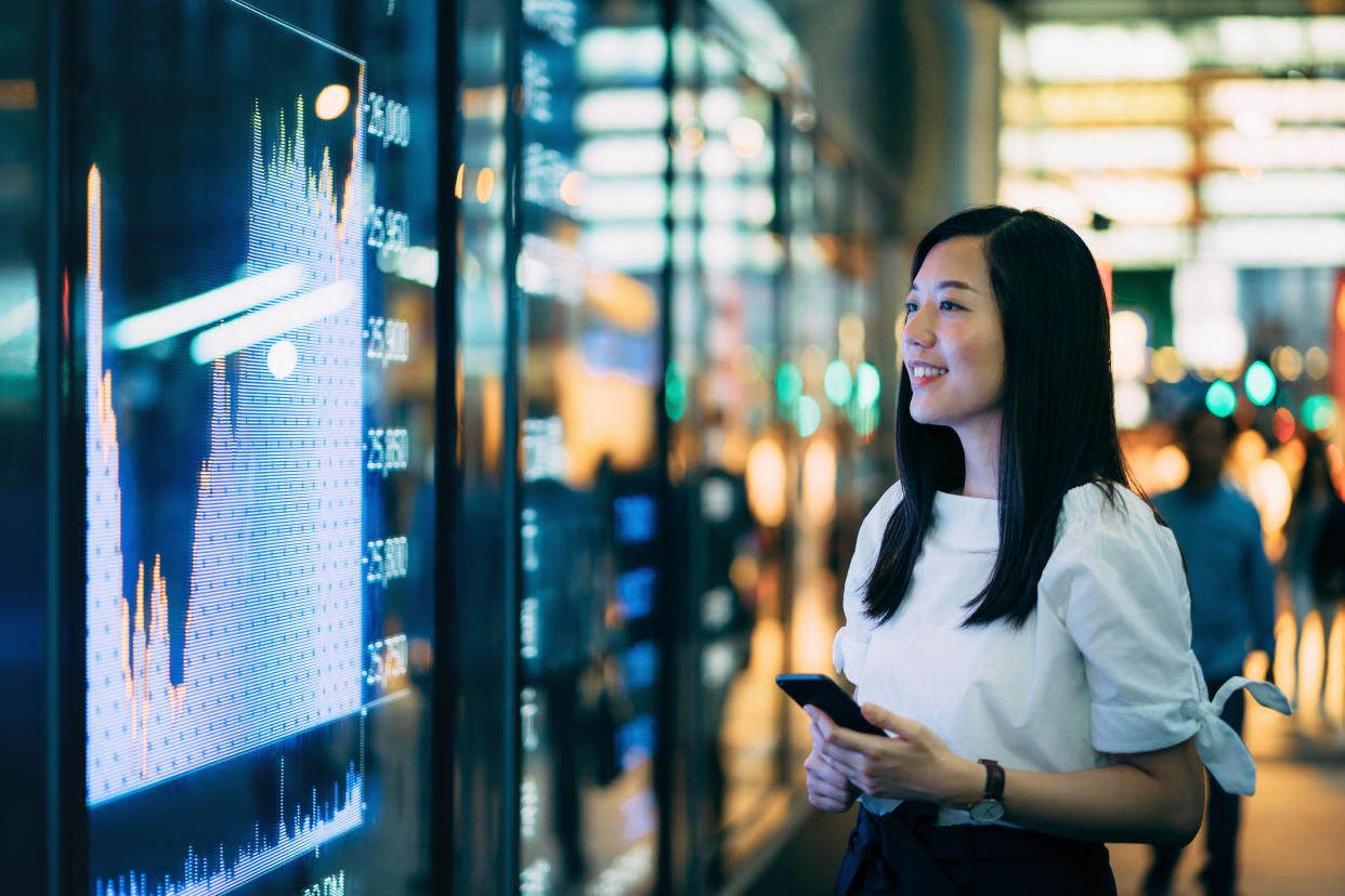 Confidence young Asian businesswoman checking financial trading data on smartphone by the stock exchange market display screen board in downtown financial district