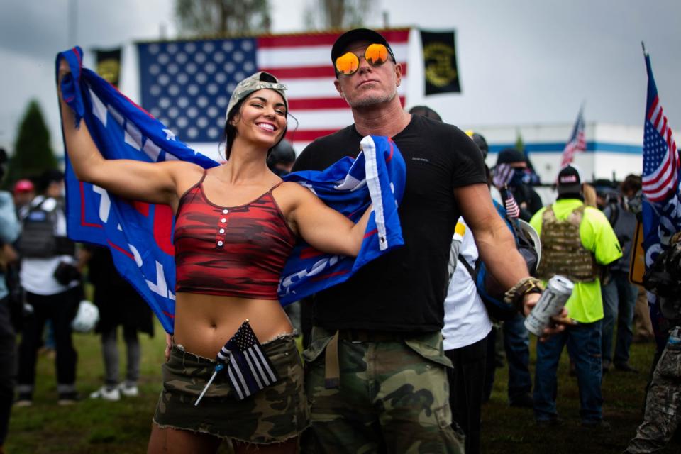 A couple wearing camo gear pose with a Trump flag at a Proud Boys rally