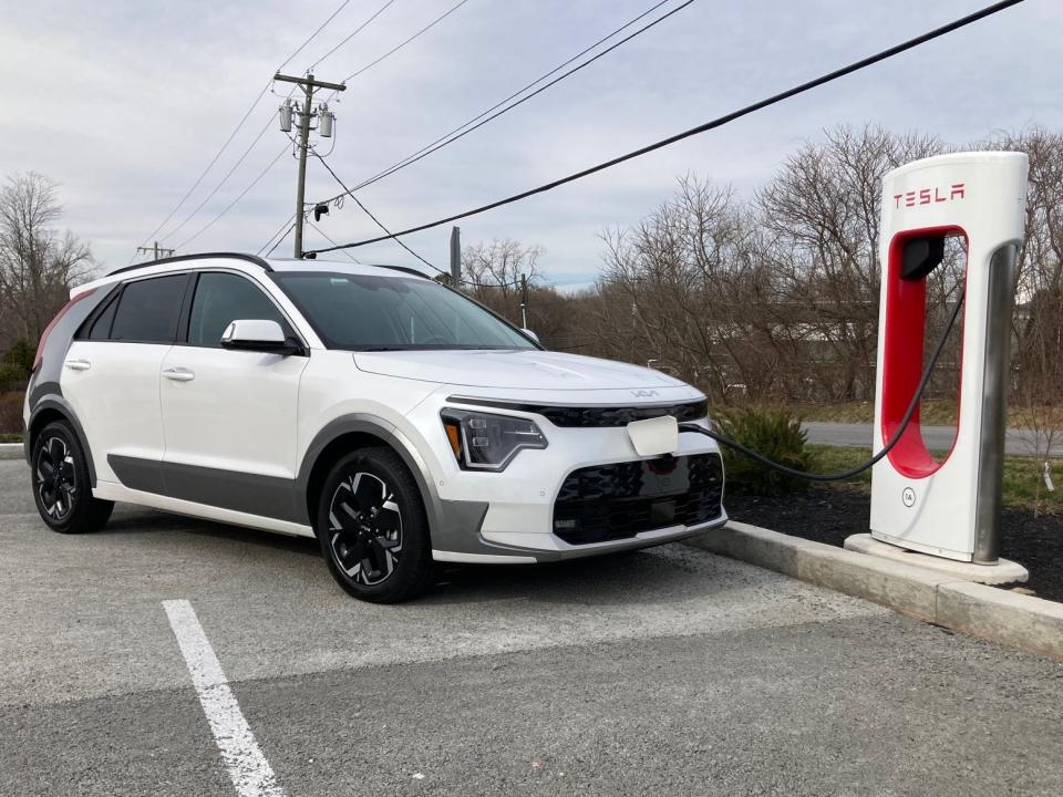 A white Kia Niro EV charges at a Tesla Supercharger in Brewster, NY, with clouds in the background.