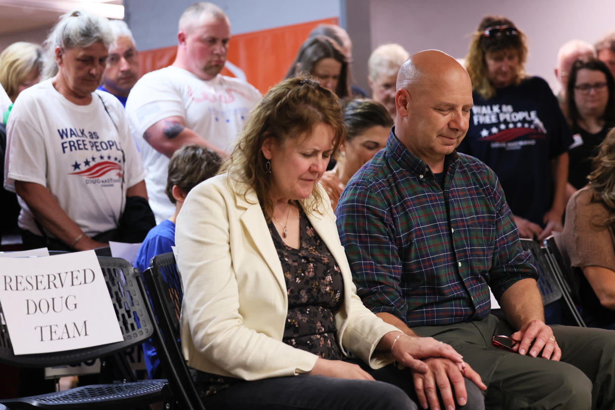 Rebecca and Doug Mastriano, seated in folding chairs, bow their heads, along with other people sitting and standing around them.