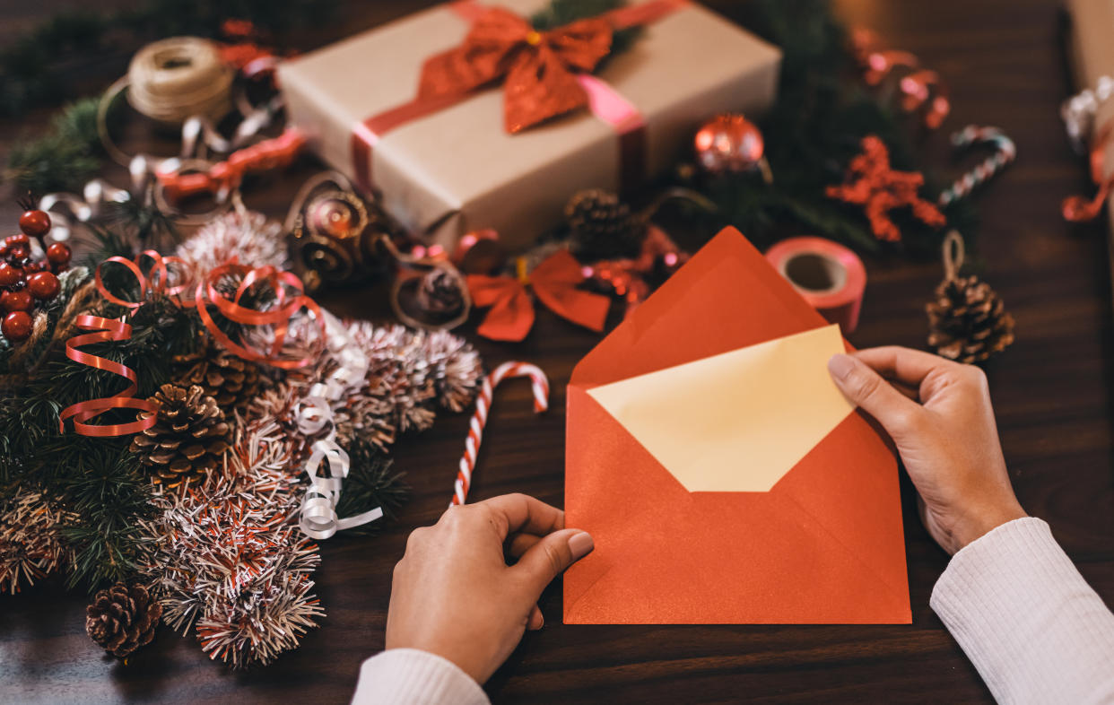 Hands removing a Christmas card from an orange envelope amid a background of holiday presents and winter evergreens and ribbon