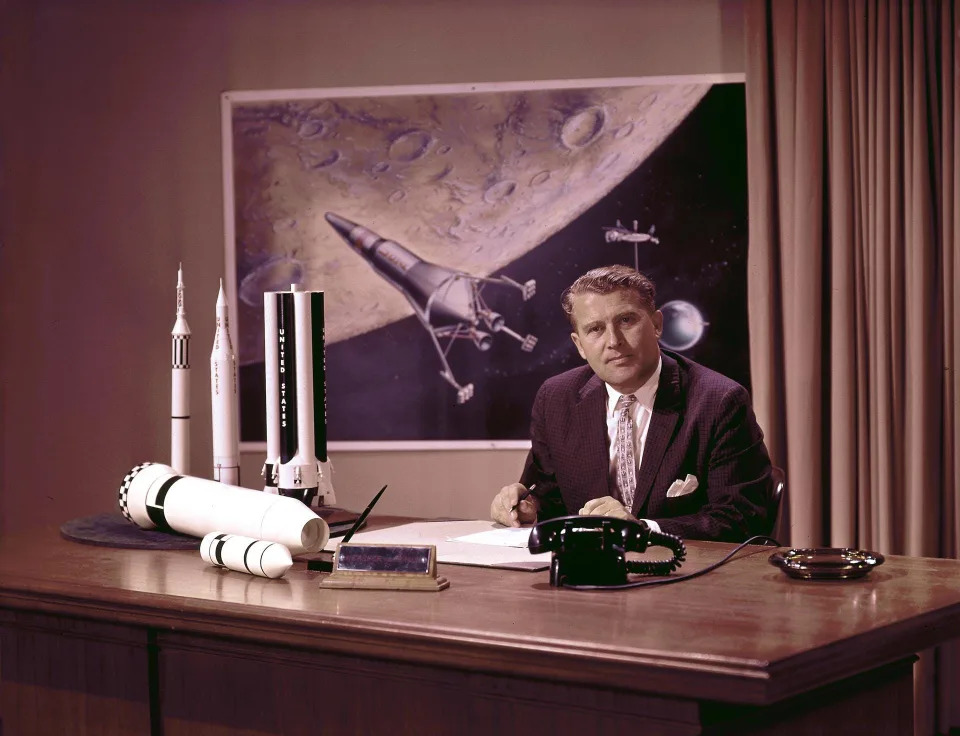 Wernher von Braun at his desk with moon lander in background and rocket models on his desk