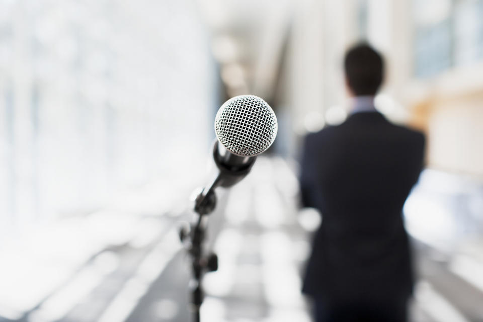 A microphone on a stand in the foreground. A man in a suit in the background.