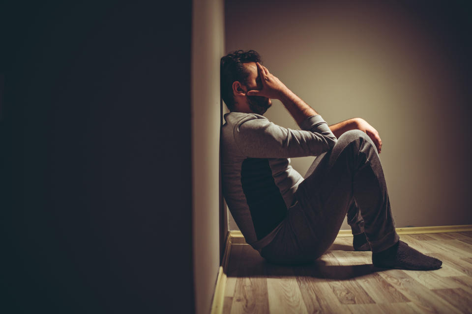 A person sits on the floor with their back against a wall, head resting on one hand, appearing distressed or in deep thought