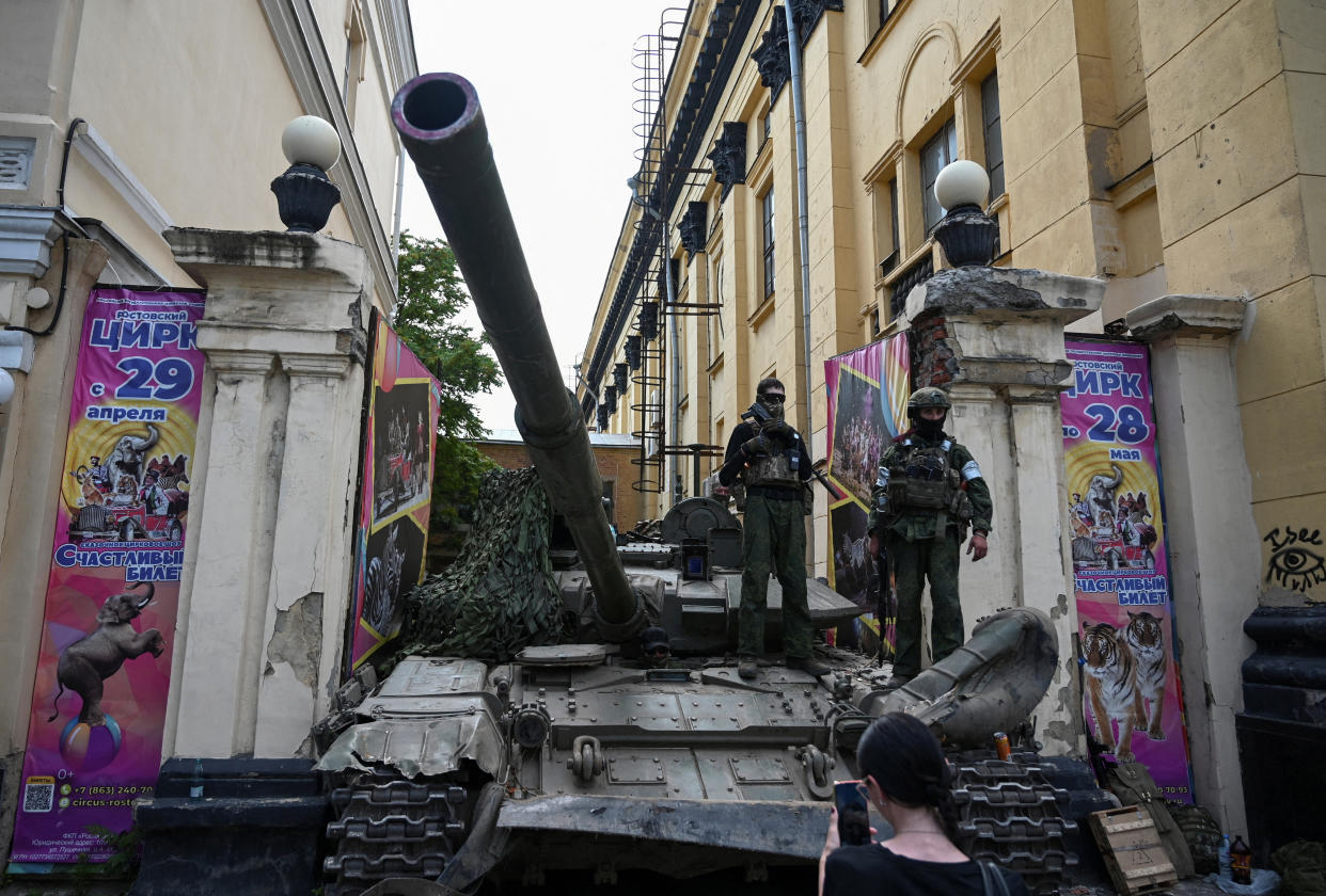 Soldiers with body armor and face masks stand atop a tank near a building between Russian language banners that appear to advertise a circus performance.