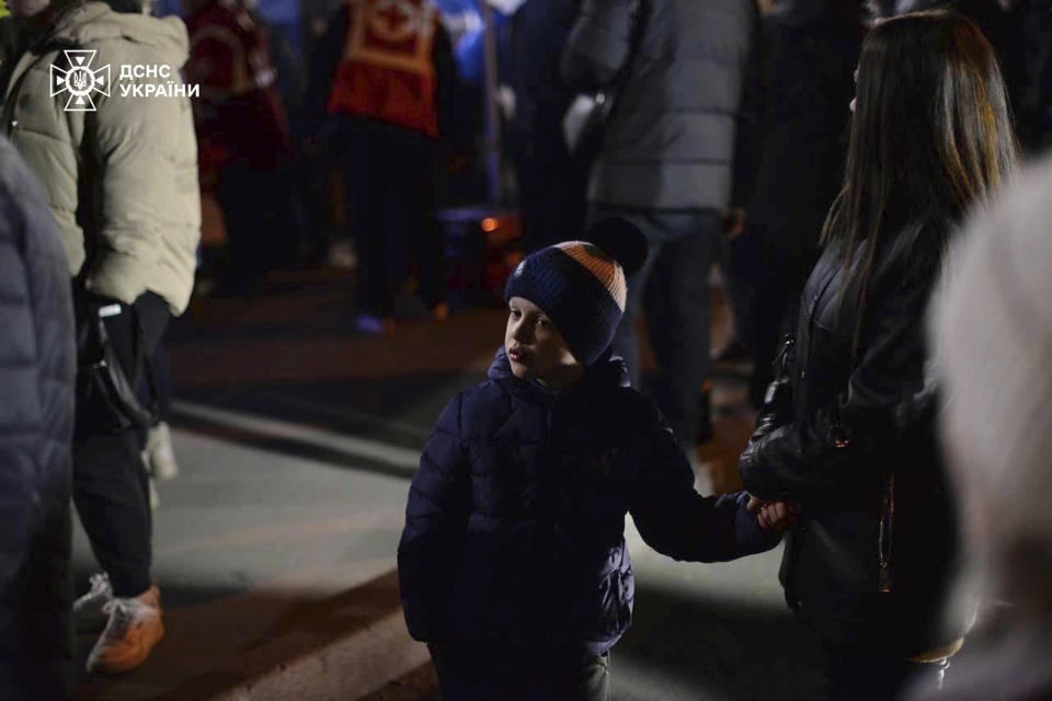 In this photo provided by the Ukrainian Emergency Service, a woman with a child stand in a yard after evacuation from an apartment building destroyed by a Russian attack in Kryvyi Rih, Ukraine, Wednesday, March 13, 2024. A few people were killed and dozens more were wounded on Tuesday evening as the result of a Russian missile strike on Zelenskyy's hometown. (Ukrainian Emergency Service via AP)