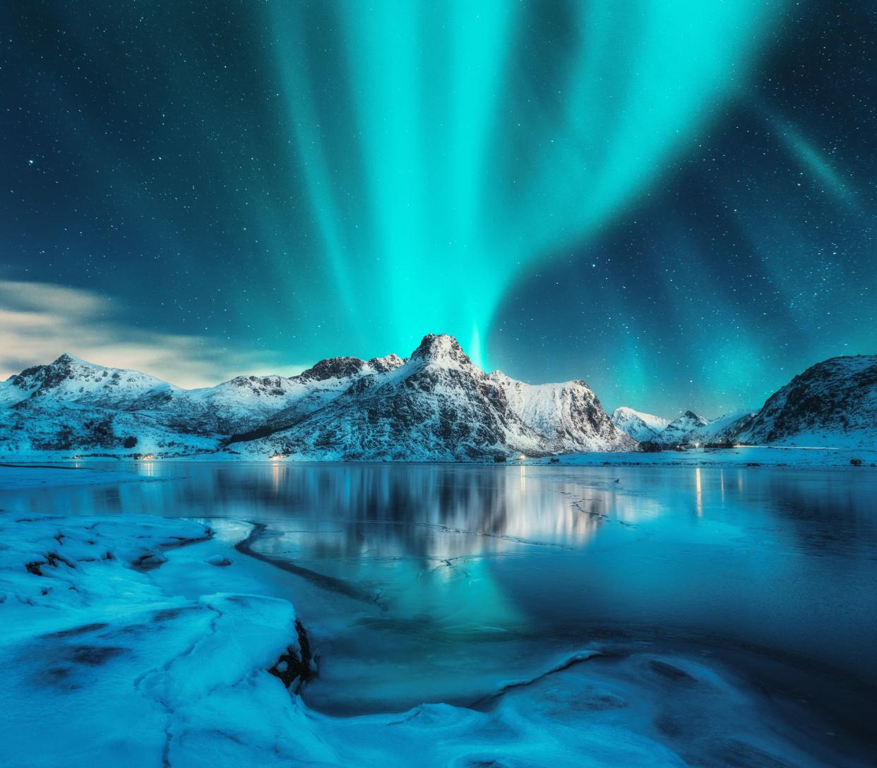 Aurora borealis over snowy mountains, frozen sea coast, reflection in water at night. Lofoten islands, Norway. Northern lights. Winter landscape with polar lights, ice in water. Starry sky with aurora