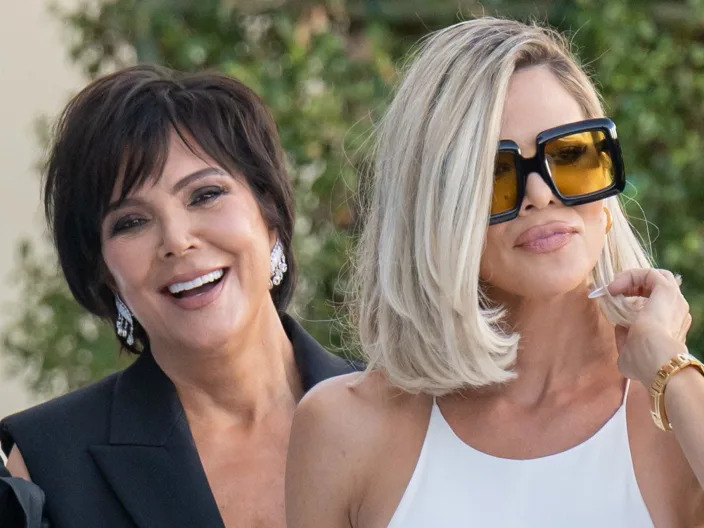 kris jenner, wearing a black suit, peeking out from behind her daughter khloe kardashian, who is wearing all white and chunky black and yellow sunglasses