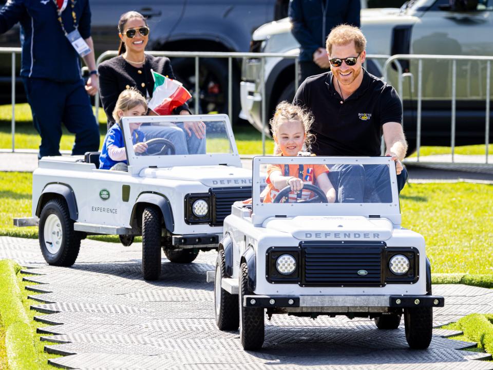 Meghan and Harry smiling and riding in child-size cars on a race track with children sitting alongside them.