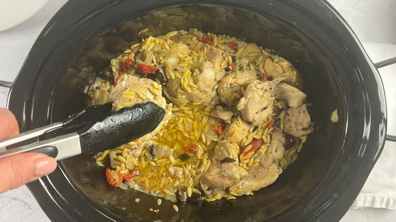 hand holding chicken with tongs over pot of orzo