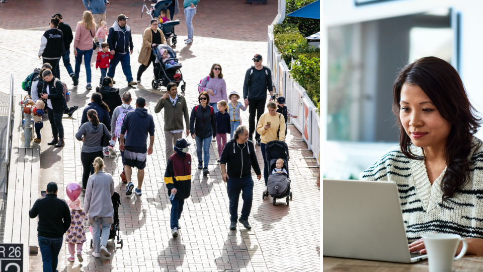 A crowd of people walk on the street and a woman works from home on her laptop.