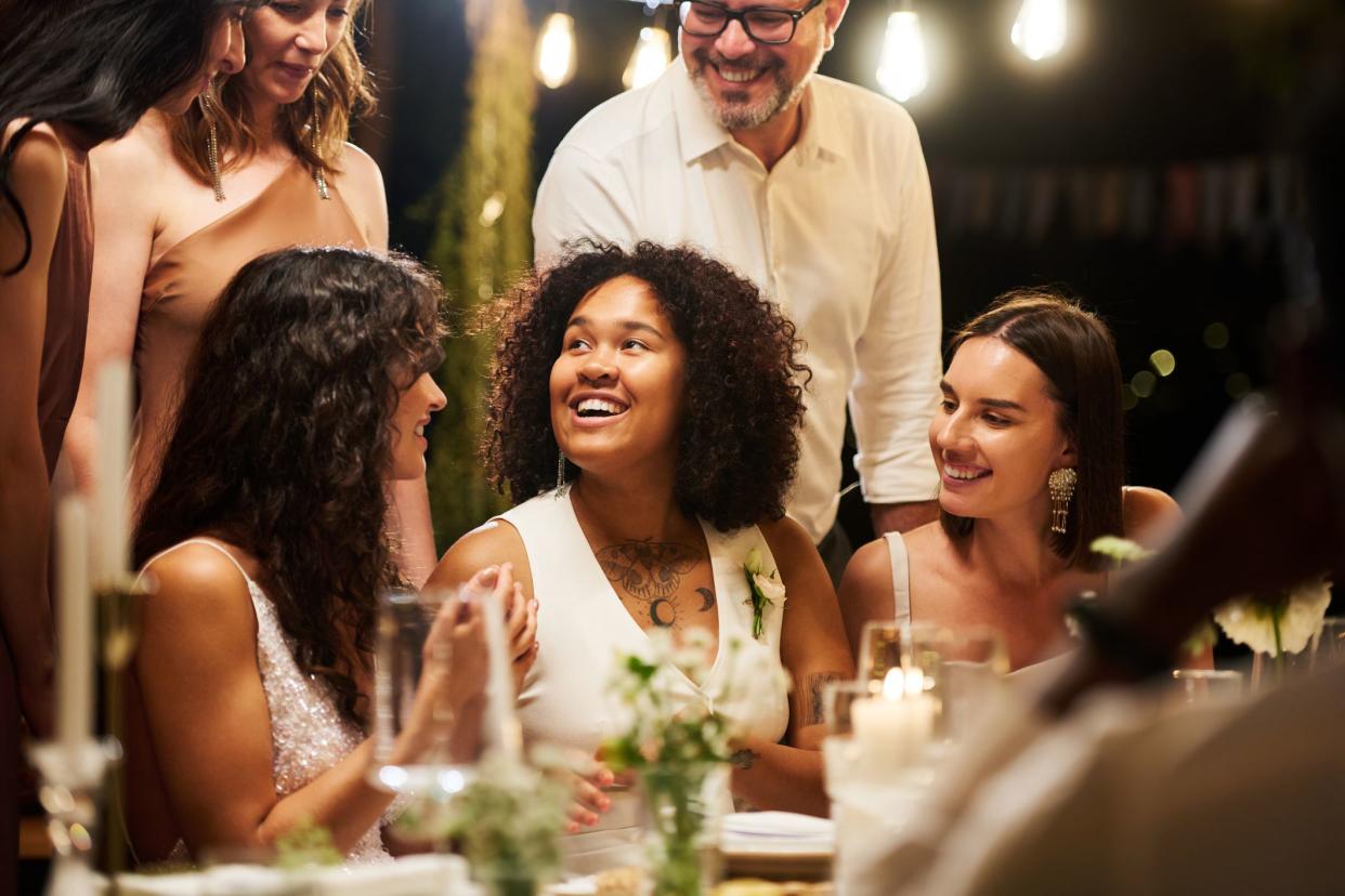 Happy young African American bride sitting by served table in restaurant among friends and wedding guests and talking to them